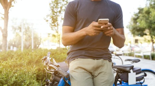 man holding smartphone leaning on bicycle during daytime